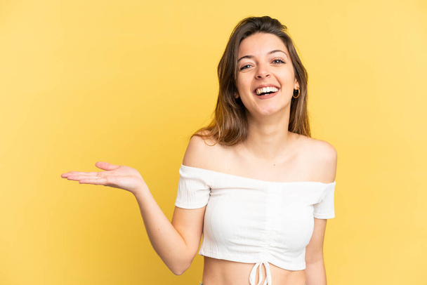 Young caucasian woman isolated on yellow background holding copyspace imaginary on the palm to insert an ad - Photo, Image