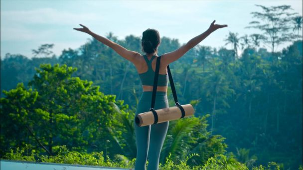 Um jovem atraente europeu morena esportes menina vai com um tapete de ioga na natureza com uma floresta tropical verde em um fundo iluminado à beira da piscina. - Foto, Imagem