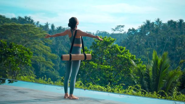 A young attractive European brunette sports girl goes with a yoga rug in nature with a green rainforest on a lit background by the pool. - Фото, зображення