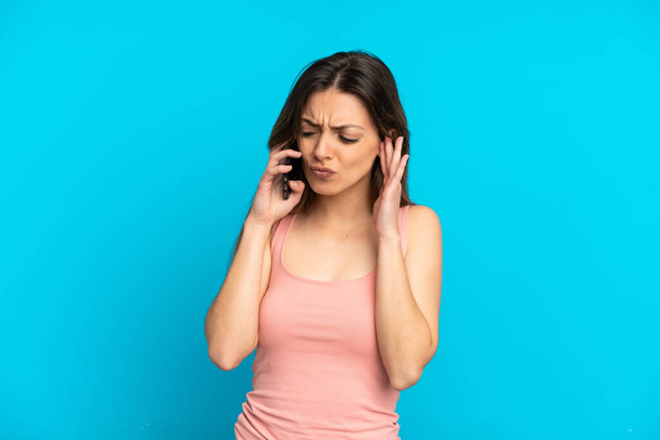 Молода біла жінка за допомогою мобільного телефону ізольована на синьому фоні розчарована і прикриває вуха
 - Фото, зображення