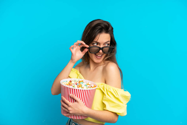 Young caucasian woman isolated on blue background with 3d glasses and holding a big bucket of popcorns - Zdjęcie, obraz