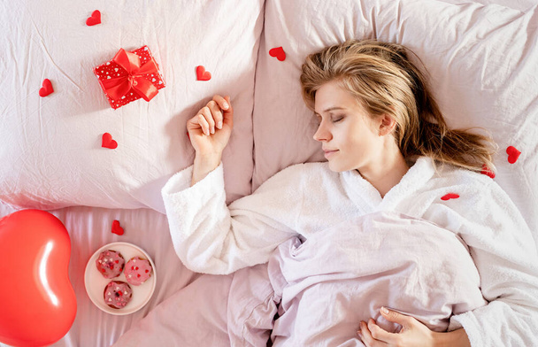 young blond woman lying in bed with holiday gift and red confetti - Foto, Imagen