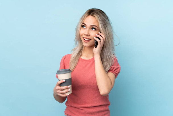 Teenager Mädchen über isoliertem blauem Hintergrund mit Kaffee zum Mitnehmen und einem Handy - Foto, Bild