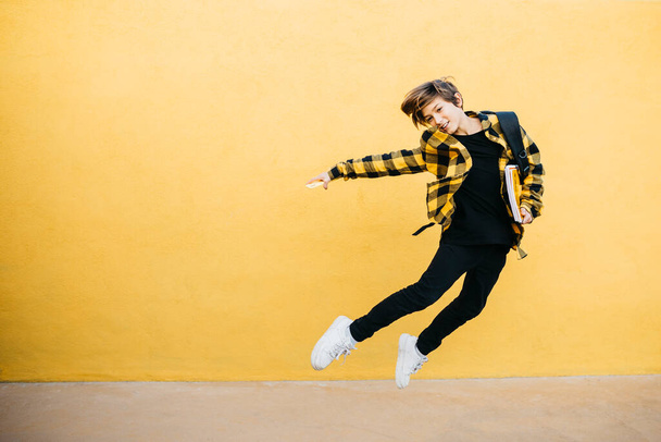 Teenager smiling student boy with notebooks and black backpack jumping in front of a yellow background - Foto, Imagem