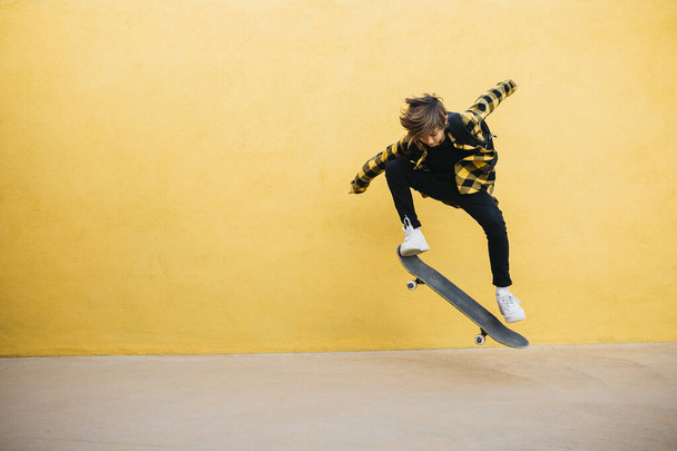 Πριν έφηβος μαθητής αγόρι άλμα με ένα skateboard μπροστά από ένα κίτρινο φόντο  - Φωτογραφία, εικόνα