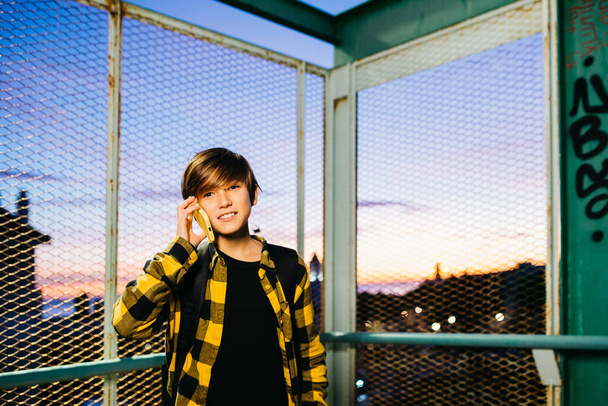 Teenager boy talking on a yellow smartphone while crossing an old metal bridge, on his way to school - 写真・画像