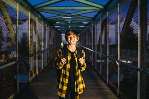 Portrait of a smiling teenager student boy crossing an old metal bridge on his way to school - Photo, Image