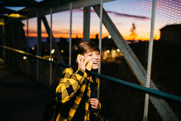 Adolescente niño hablando en un teléfono inteligente amarillo mientras cruza un viejo puente de metal, en su camino a la escuela - Foto, imagen