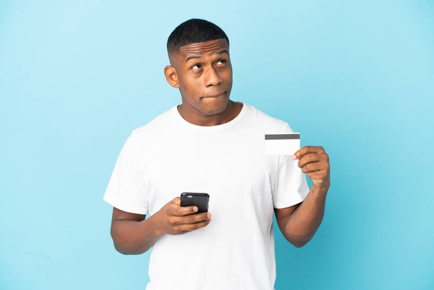 młody Latynos człowiek odizolowany na niebieskim tle zakup z telefonu komórkowego z karty kredytowej podczas myślenia - Zdjęcie, obraz