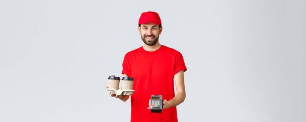 Food delivery, quarantine, stay home and order online concept. Friendly bearded courier in red uniform handing coffee delivery and POS terminal for client to pay contactless, grey background - Photo, Image