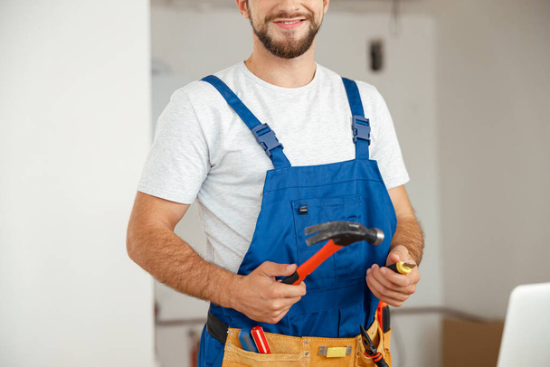 Обрізаний знімок професійного майстра в уніформі посміхається на камеру і тримає молоток під час ремонту в приміщенні
 - Фото, зображення