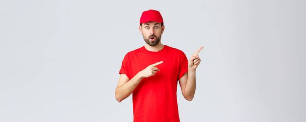 Online shopping, delivery during quarantine and takeaway concept. Take quick look here. Impressed courier showing clients special promo. Employee in red uniform pointing fingers right - Photo, Image