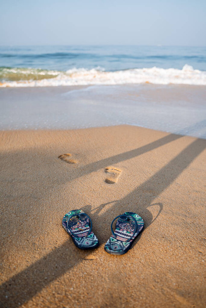 Blue beach slippers on a sandy beach. Footprints of a man on the shore. Rest by the sea, summer adventures. Surf noise and sea foam - Fotó, kép