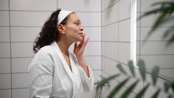 Side view of a charming middle aged African pretty woman applying cleansing exfoliating beauty product on her face, looking at her reflection in the bathroom mirror. Facial skin care concept - Imágenes, Vídeo