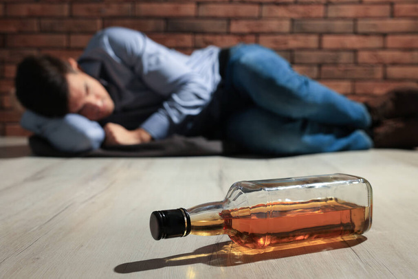 Addicted man lying near red brick wall, focus on bottle of alcoholic drink - Fotó, kép