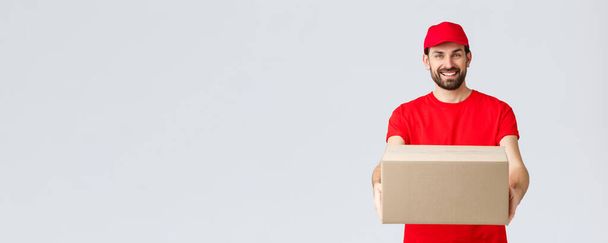 Order delivery, online shopping and package shipping concept. Friendly smiling bearded courier in red uniform t-shirt and cap, giving out package for customers, handing box to client - Photo, Image