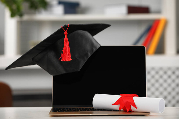 Graduation hat, student's diploma and laptop on white table indoors - Photo, image