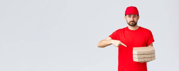 Food delivery, quarantine, stay home and order online concept. Confident friendly courier in red uniform cap and t-shirt, employee bring order pizza, pointing finger at boxes, grey background - Photo, Image