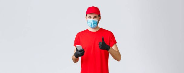 Contactless delivery, payment and online shopping during covid-19, self-quarantine. Friendly courier in gloves and red uniform cap, face mask, show thumb-up as recommend smartphone app or site - Photo, Image