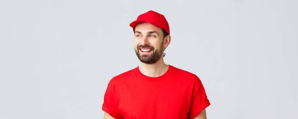 オンラインショッピング、隔離中の配達とテイクアウトの概念。明るい笑顔で離れて見て、赤い制服キャップとTシャツの陽気なひげを生やした男、バナーサインを読んで、灰色の背景 - 写真・画像