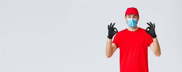 Ковид-19, самокарантин, онлайн-шопинг и концепция доставки. Возбужденный курьер в красной кепке, футболке и маске для лица, работающий над вспышкой коронавируса, показывающий признаки безопасности, гарантирующий курьерскую службу
 - Фото, изображение