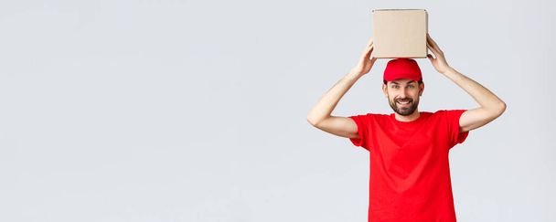注文配達、オンラインショッピングとパッケージ配送の概念。赤の制服キャップとTシャツの面白いとかわいい髭の宅配便は、頭の上に箱を保持。笑顔のパッケージの従業員 - 写真・画像