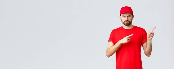 Online nakupování, doručování během karantény a donášky konceptu. Rozzlobený kurýr v červené uniformě s čepicí a tričkem, zamračený mrzutý, nesouhlasně ukazující prsty, se cítí znepokojen - Fotografie, Obrázek