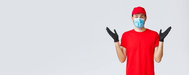 Groceries and packages delivery, covid-19, quarantine and shopping concept. Happy rejoicing delivery guy in red uniform, face mask and gloves receive good news, clap hands applause surprised - Photo, Image
