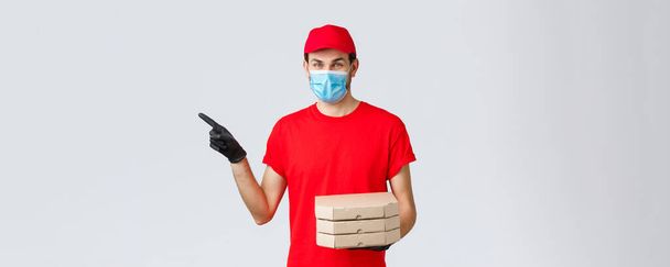 Food delivery, application, online contactless shopping and covid-19 concept. Charismatic courier in red uniform, face mask and gloves, pointing finger left, holding pizza, bring order client - Fotoğraf, Görsel