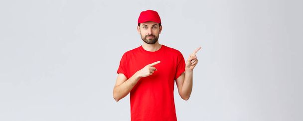 オンラインショッピング、隔離中の配達とテイクアウトの概念。赤い制服キャップとTシャツに自信を持って髭を生やした宅配便,笑顔のカメラ,方向を与える,指の右を指して,クライアントに通知 - 写真・画像