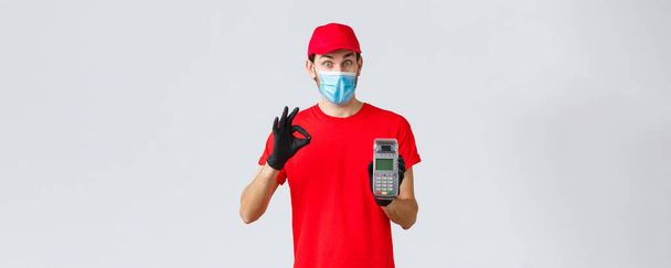 Contactless delivery, payment and online shopping during covid-19, self-quarantine. Excited courier in red uniform, face mask and gloves recommend pay order with POS terminal and credit card - Φωτογραφία, εικόνα