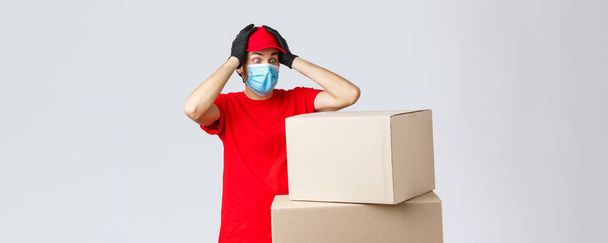 Packages and parcels delivery, covid-19 quarantine and transfer orders. Concerned and troubled courier in red uniform, face mask and gloves, grab head and gasping shocked staring at boxes - Photo, Image