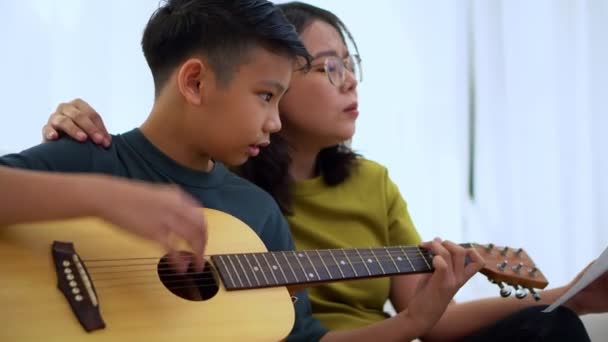 Asyalı anne, oğlu ve gitar çalan Asyalı çocuğu kucaklıyor ve annesi kanepede sarılıyor ve takdir edilip cesaretlendirildiğini hissediyor. Mutlu bir aile kavramı, öğrenme ve eğlenceli yaşam tarzı, aile bağlarını sevme - Video, Çekim