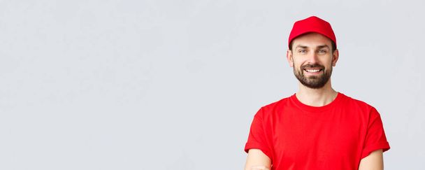 Online shopping, delivery during quarantine and takeaway concept. Handsome bearded courier in red uniform, smiling cheerful, cross arms chest, ready to deliver your order, grey background - Fotografie, Obrázek