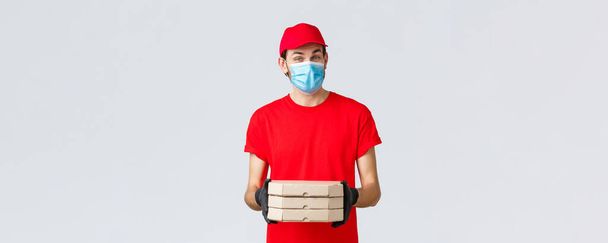 Food delivery, application, online grocery, contactless shopping and covid-19 concept. Pleasant courier in red uniform bring pizza in boxes, wear face mask and rubber gloves, give clint order - Foto, Bild