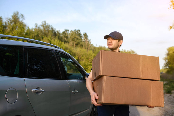 Young man courier holding a paper box, behind him a background a car - Foto, imagen