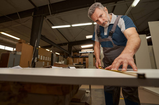 Concentrated senior male person measuring wooden plank - Foto, Bild