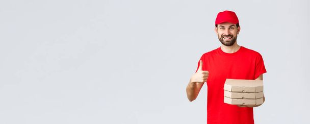 Food delivery, quarantine, stay home and order online concept. Friendly smiling bearded courier in red uniform cap and t-shirt, recommend their service or restaurant, give pizza to customer - Photo, Image