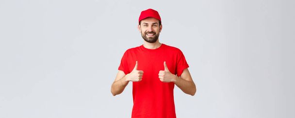 Online shopping, delivery during quarantine and takeaway concept. Cheerful courier in red uniform cap and t-shirt, recommends make orders, thumbs-up in approval, grey background - Photo, Image