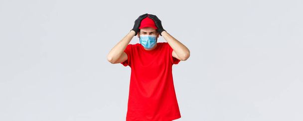 Covid-19, self-quarantine, online shopping and shipping concept. Troubled and embarrassed courier man in medical mask, gloves, hold hands on head, grimacing, making mistake with parcel delivery - Photo, Image