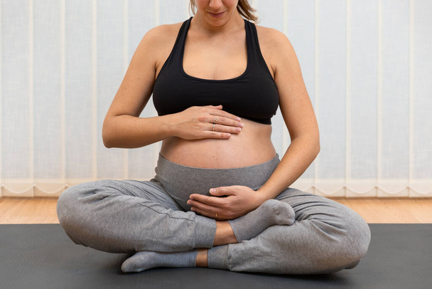 Zrelaksowana ciężarna kobieta siedzi na macie i ćwiczy jogę dotykając brzucha w centrum zdrowia - Zdjęcie, obraz