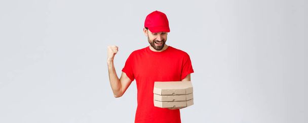 食料の配達、隔離、家にいてオンラインの概念を注文しなさい。喜んでピザの注文を見て灰色の背景に赤いTシャツとキャップの幸せな興奮したひげそりの宅配業者 - 写真・画像