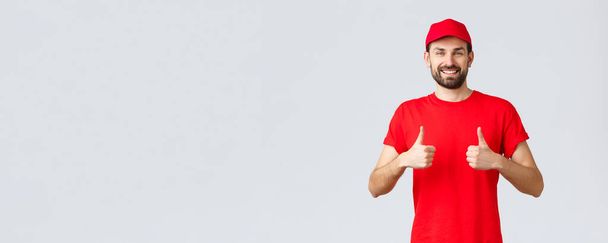 Online shopping, delivery during quarantine and takeaway concept. Cheerful courier in red uniform cap and t-shirt, recommends make orders, thumbs-up in approval, grey background - Photo, Image