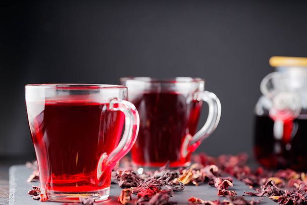 Hibiscus tea in glass mug on a dark background. Cup of red hibiscus tea and dry hibiscus petals on a slate board - Fotó, kép
