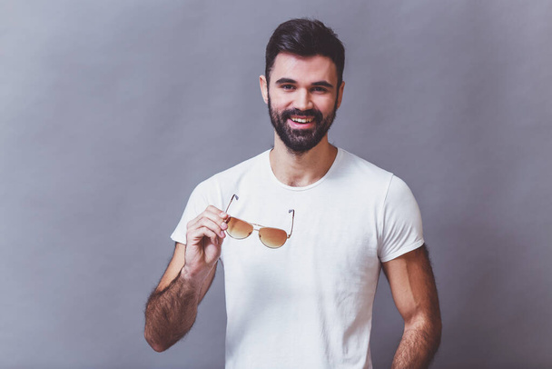 Portrait d'un beau jeune homme avec une barbe, un t-shirt blanc, garde les lunettes à la main, souriant, regardant dans la caméra, sur un fond gris
 - Photo, image