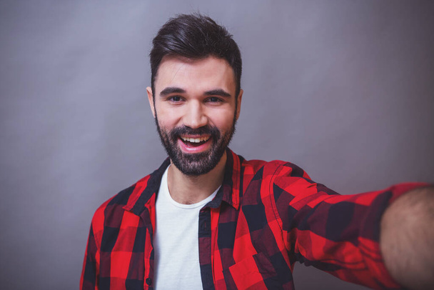 Красивий молодий чоловік з бородою в картатій сорочці робить себе, посміхаючись, на сірому фоні
 - Фото, зображення