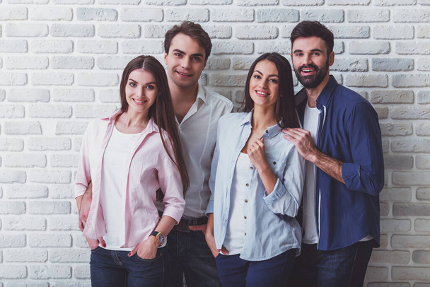 Groep van vier jonge mooie mensen in alledaagse kleren, poseren in de Studio, glimlachend, kijkend naar de camera, tegen een bakstenen muur - Foto, afbeelding