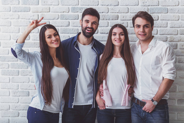 Group of four young beautiful people in everyday clothes,  posing in the studio, smiling, looking at the camera, against a brick wall - Photo, image