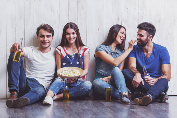 Four attractive young friends sitting on the floor, keeping beer and popcorn, smiling - Фото, изображение
