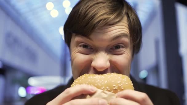 Portret van een jongeman die een hamburger bijt en naar de camera kijkt, close-up - Video